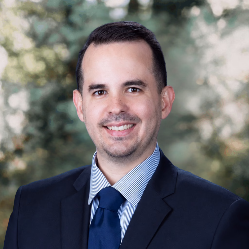 Smiling man in a navy suit and blue tie, outdoor headshot with blurred greenery.